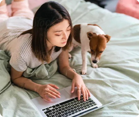 Woman laying on a bed looking at a laptop with a terrier dog at her side.