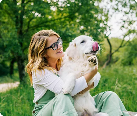 A female pet owner with glasses sitting outdoors with a white dog in her lap licking its nose.