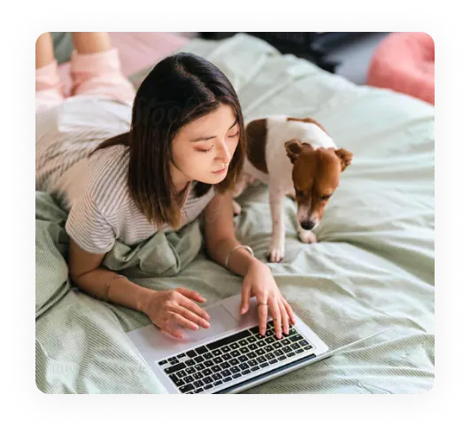 Woman laying on a bed looking at a laptop with a terrier dog at her side.